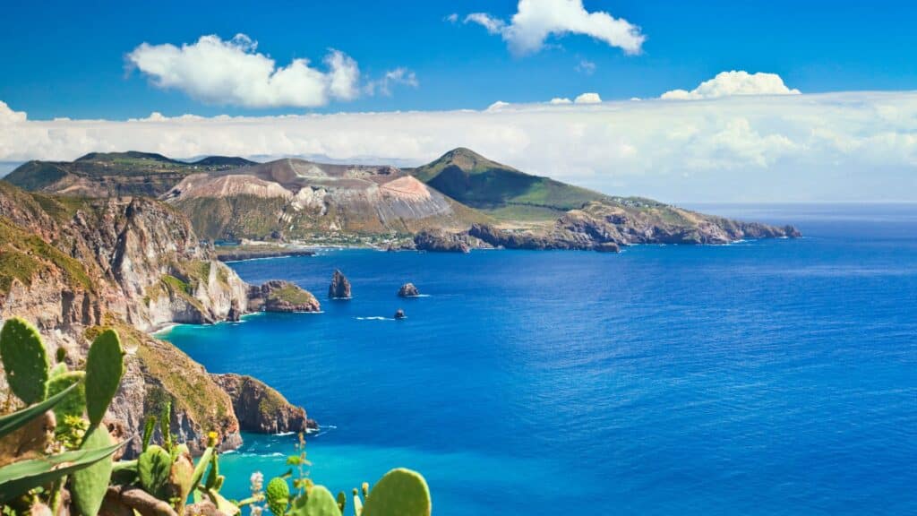 A panoramic view of the Aeolian Islands' coastline, featuring rugged cliffs, crystal-clear blue waters, and fertile volcanic land, illustrating the unique terroir of Sicilian vineyards.