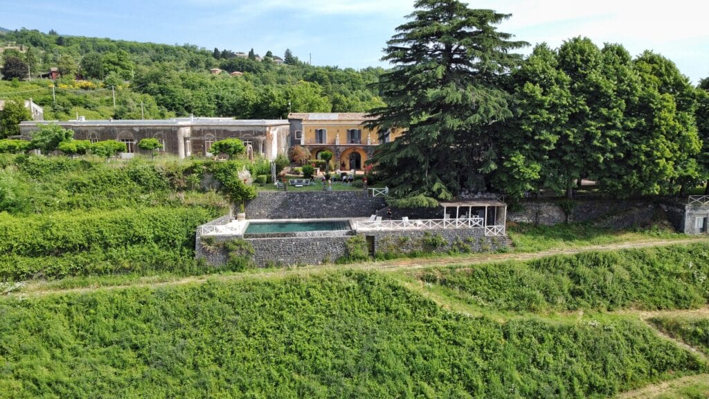 A scenic view of the Barone di Villagrande vineyard in Sicily, featuring its traditional winery structure and a serene swimming pool, surrounded by lush grapevines that embody the spirit of sustainable travel and cultural immersion.