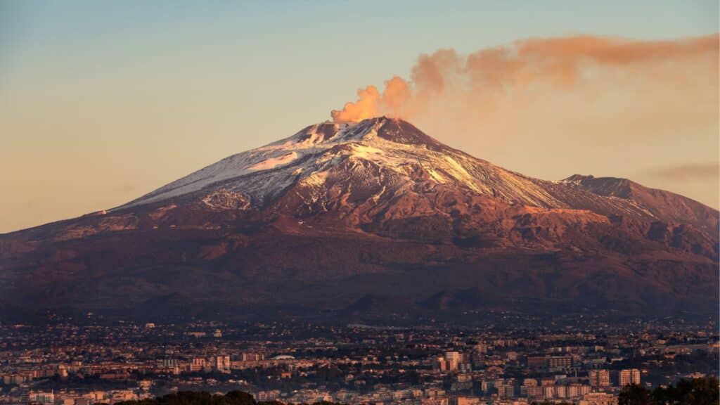 Mount Etna with smoke rising from its peak, seen from a distance with the city of Catania in the foreground, illustrating the volcanic landscape that influences Sicilian winemaking, particularly renowned for Sicilian volcanic wines