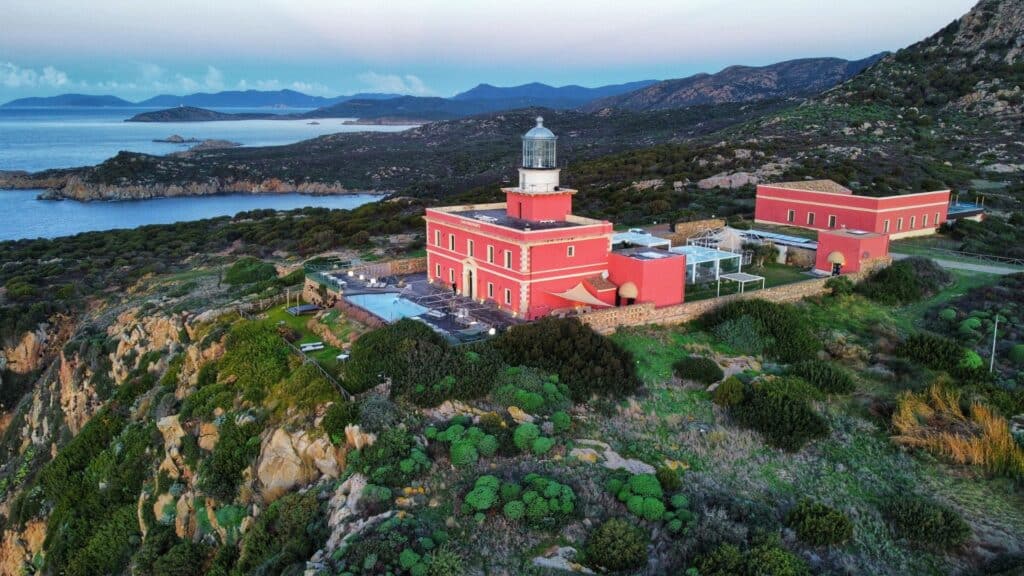 A historic red lighthouse turned boutique hotel perched on a cliffside in Sardinia, with panoramic views of the sea and surrounding rugged landscape.