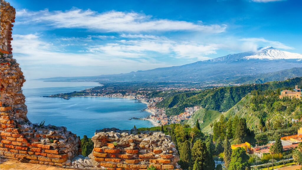 Panoramic view of Sicily’s coastline with historic ruins and the majestic Mount Etna in the background.