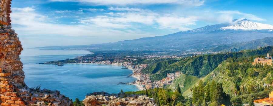 Panoramic view of Sicily’s coastline with historic ruins and the majestic Mount Etna in the background.