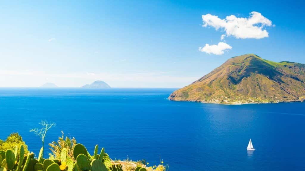 A serene view of the Aeolian Islands with crystal-clear blue waters, a sailboat, and lush green volcanic hills.