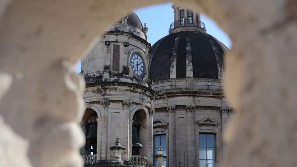 Historic domes of Catania's Piazza del Duomo viewed through a stone arch, highlighting the city’s architectural beauty.