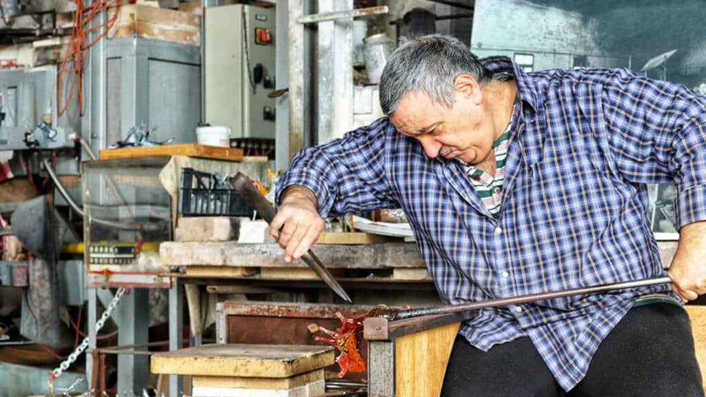 A skilled glass artisan shaping molten glass with tools in a traditional workshop, surrounded by industrial equipment and creative clutter.