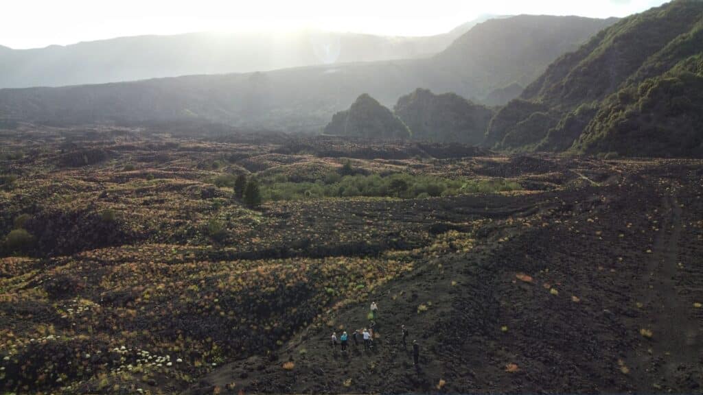 Visitors hiking along the volcanic landscape of Mount Etna with rugged terrain and surrounding hills in Sicily.