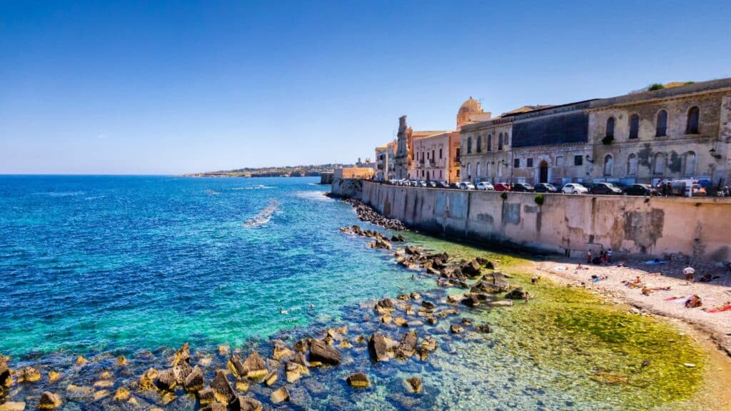 Ortigia’s coastline with crystal-clear waters and historic buildings lining the shore in Syracuse, Sicily.