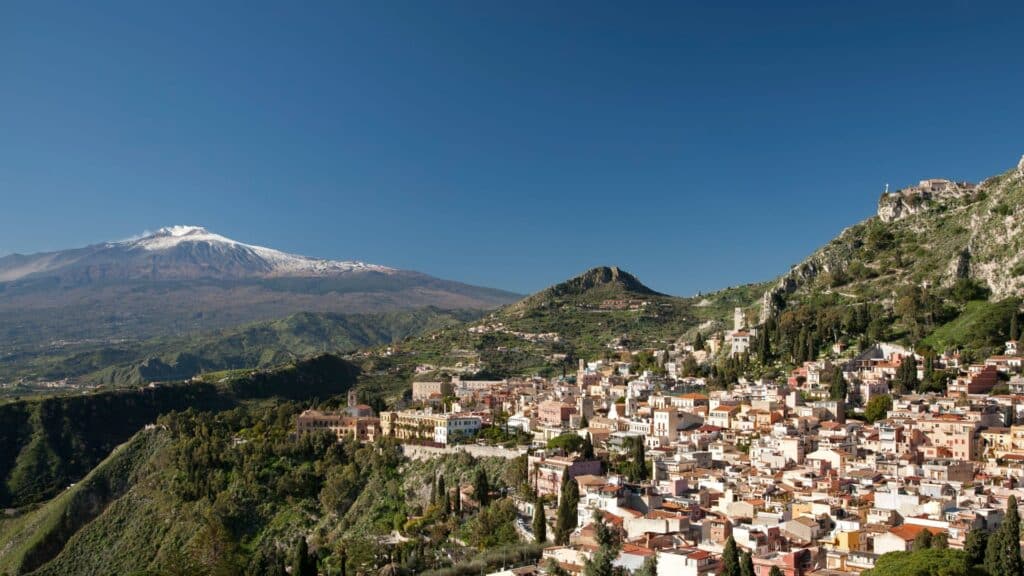 Panoramic view of Taormina, Sicily with Mount Etna in the background and colorful houses on a hill.