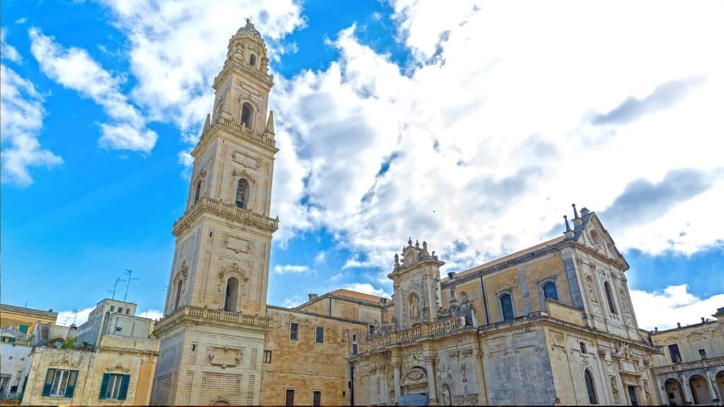 The Baroque bell tower of Lecce Cathedral in Italy, set against a bright blue sky with scattered clouds, showcasing the intricate stonework of the "Florence of the South."