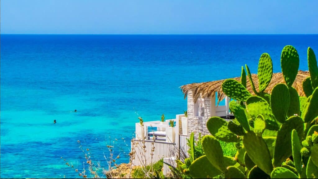 A view of the crystal-clear waters and white sand beaches of Santa Maria di Leuca, with a small white stone building and cactus plants in the foreground.