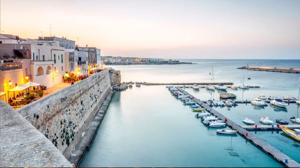 A peaceful evening in Otranto, Italy, with views of the marina, boats, and white-walled buildings lining the coast, under the soft glow of sunset.