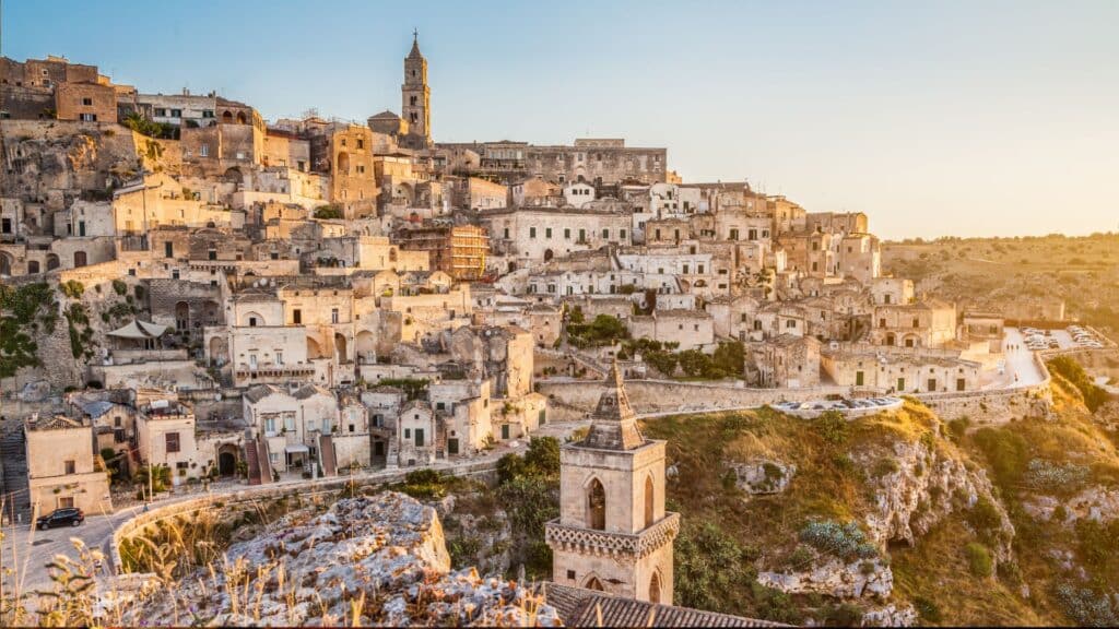 A panoramic view of Matera's ancient Sassi cave dwellings carved into rock, with restored boutique hotels and cultural spaces, under a clear blue sky.