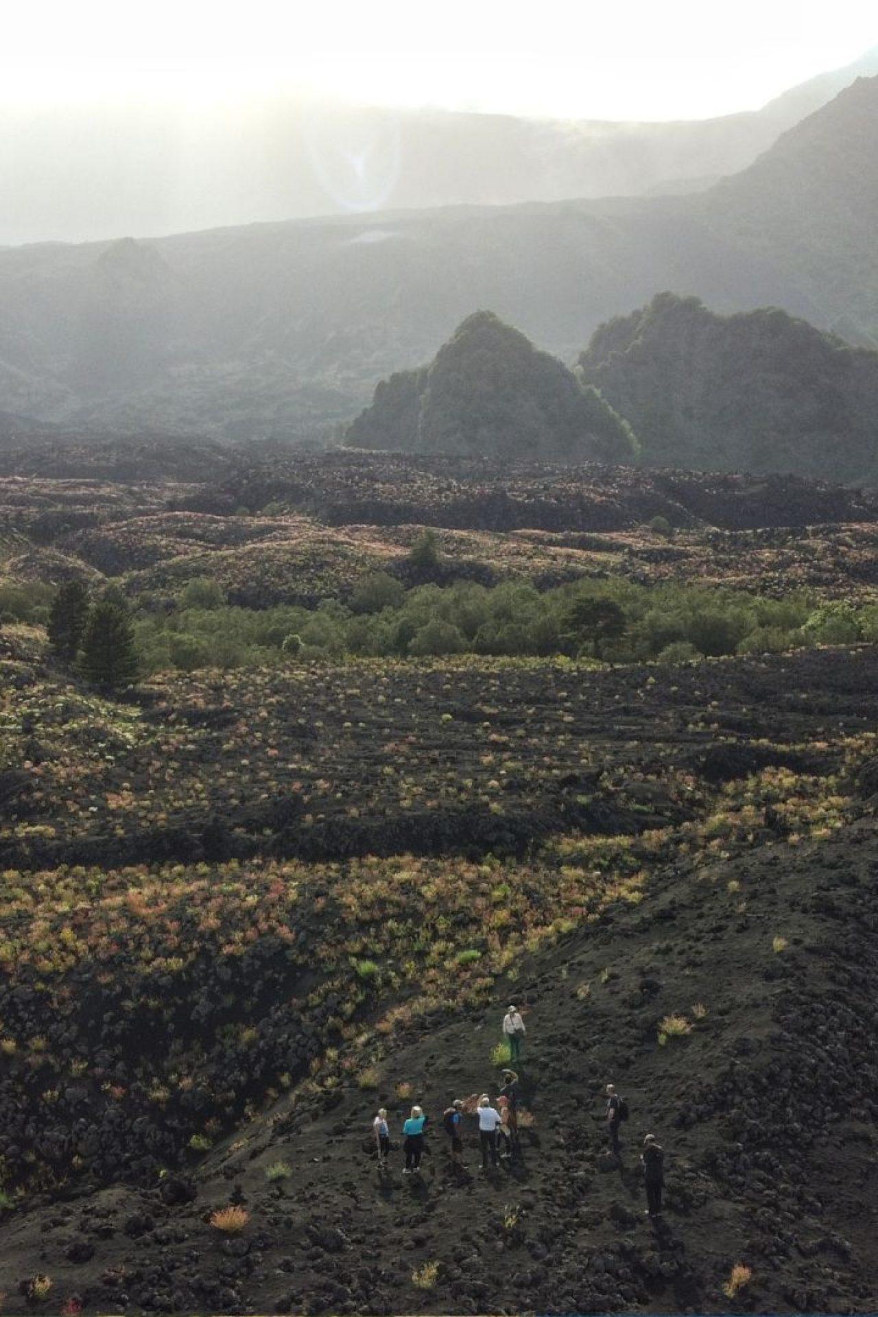 Visitors hiking along the volcanic landscape of Mount Etna with rugged terrain and surrounding hills in Sicily.